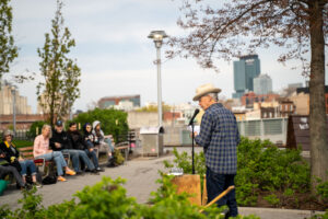 A man in a cowboy hat stands at a microphone, speaking to people seated on benches across from him. The Brooklyn skyline is in the background.