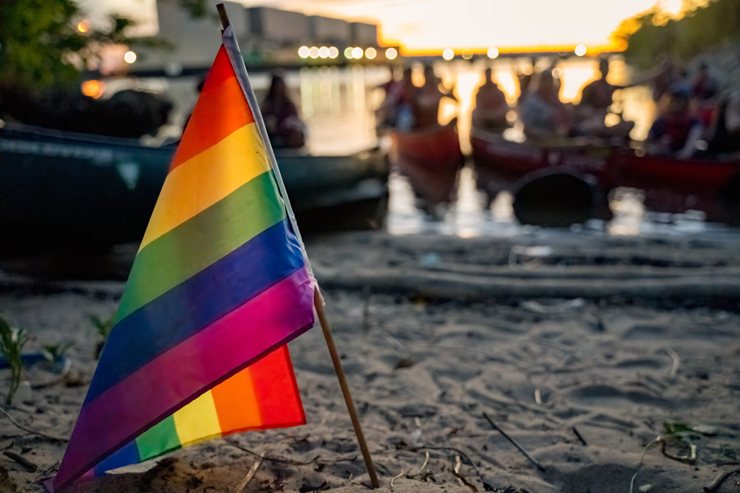 A rainbow flag stuck in the sand, canoes in the background out of focus, as the sun sets behind them.