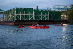 Multiple red canoes with people in them paddle in front of barges filled with green shipping containers, floating on the New York waterfront.